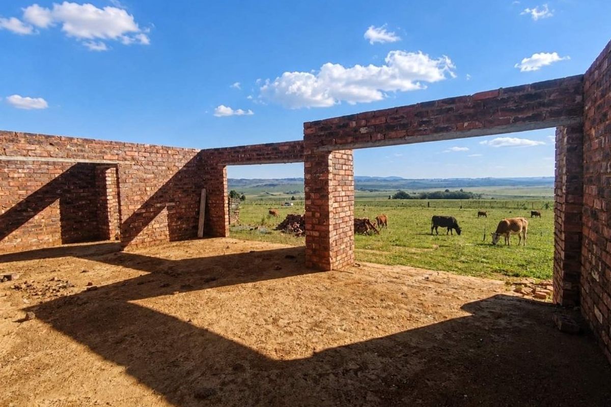 An unfinished brick structure under construction, featuring open wall frames with expansive views over the surrounding farm landscape.