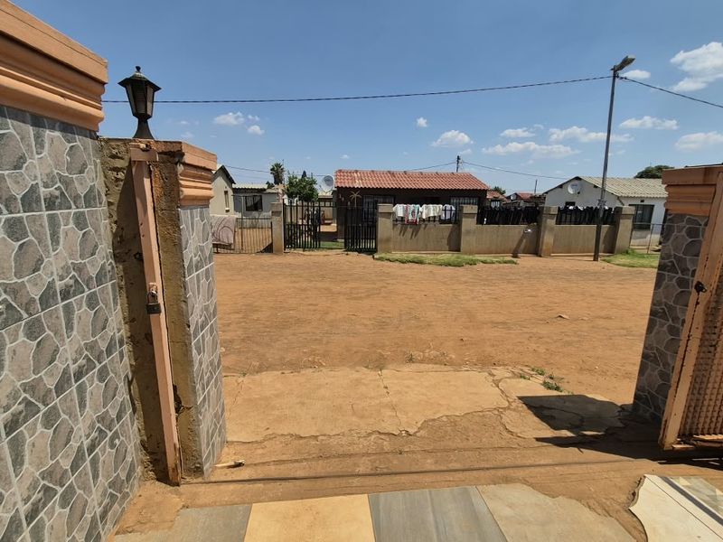 Front entrance view showing a stone-clad perimeter wall, metal gate, and an expansive unpaved driveway area leading to the main property.