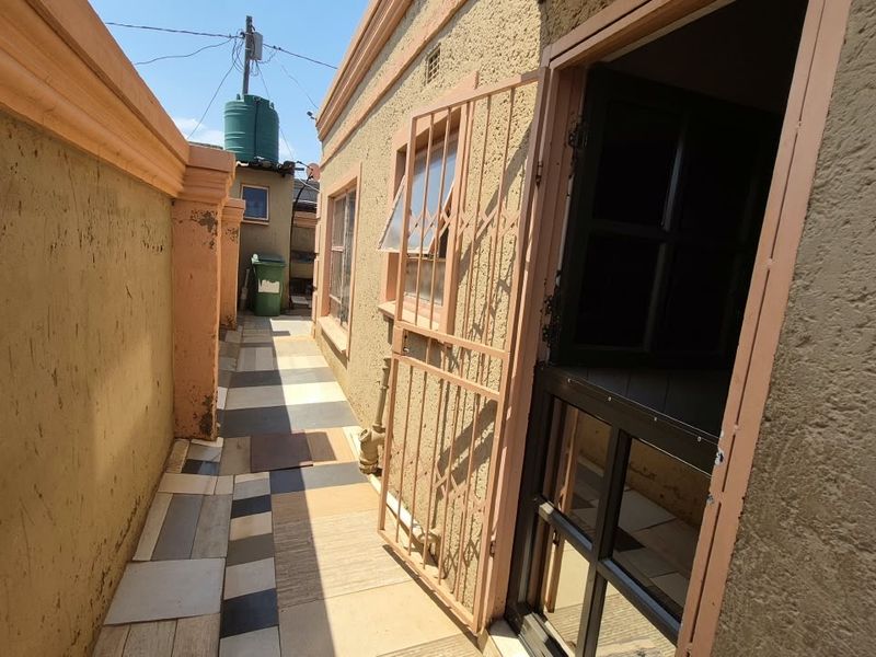 A narrow exterior side passage featuring a tiled walkway, security gate, and boundary wall with visible signs of wear.