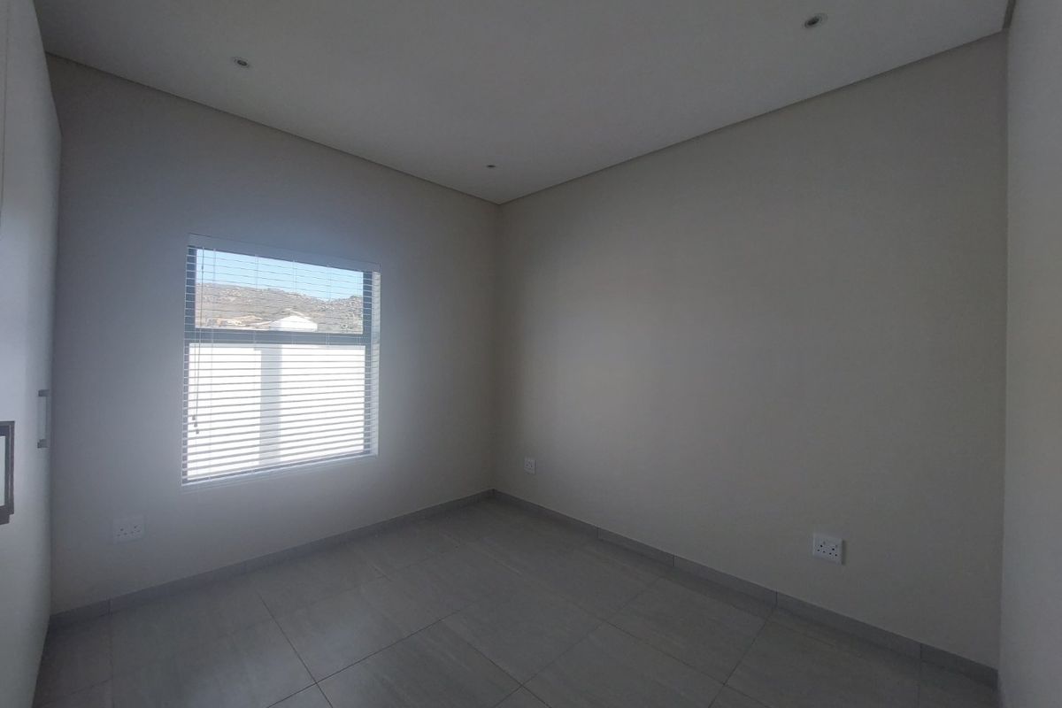 A clean, neutral-toned bedroom featuring tiled flooring, built-in cupboards, recessed lighting, and a window fitted with venetian blinds.