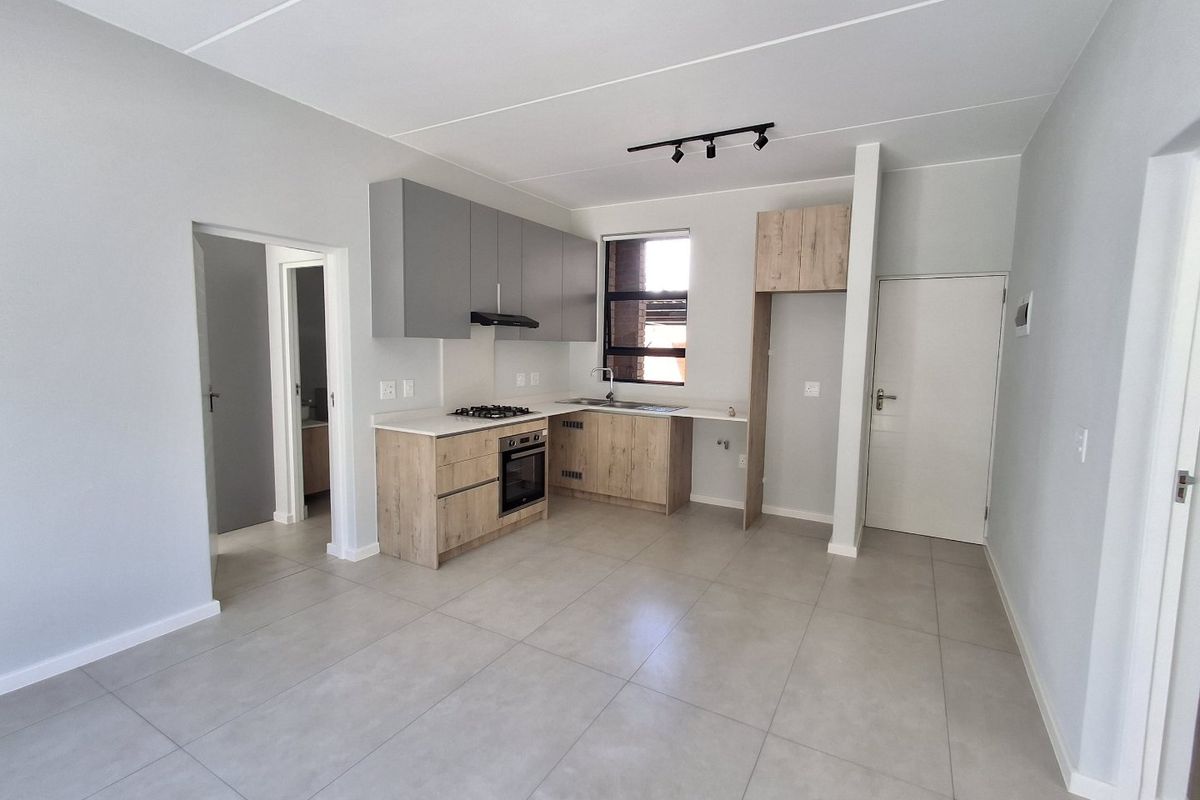 Modern open-plan kitchen featuring light wood-look cabinetry, grey upper cupboards, built-in oven and hob, and neutral floor tiles in a bright, clean space.