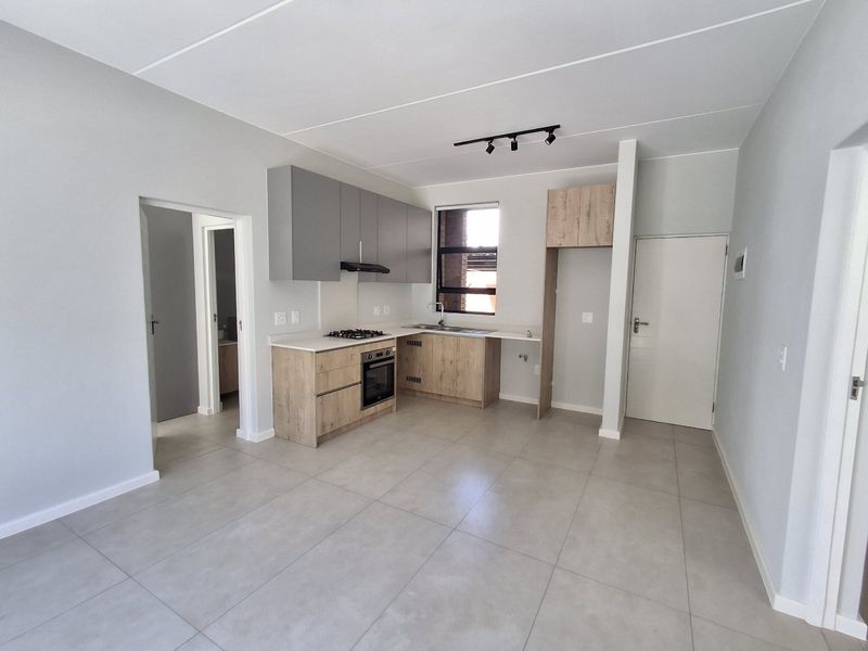 Modern open-plan kitchen featuring light wood-look cabinetry, grey upper cupboards, built-in oven and hob, and neutral floor tiles in a bright, clean space.