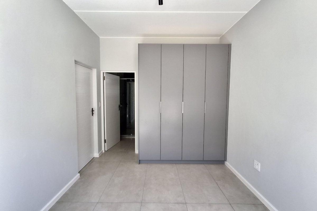 A clean, modern bedroom featuring neutral grey walls, large-format floor tiles, and ample built-in cupboard storage with a minimalist aesthetic.