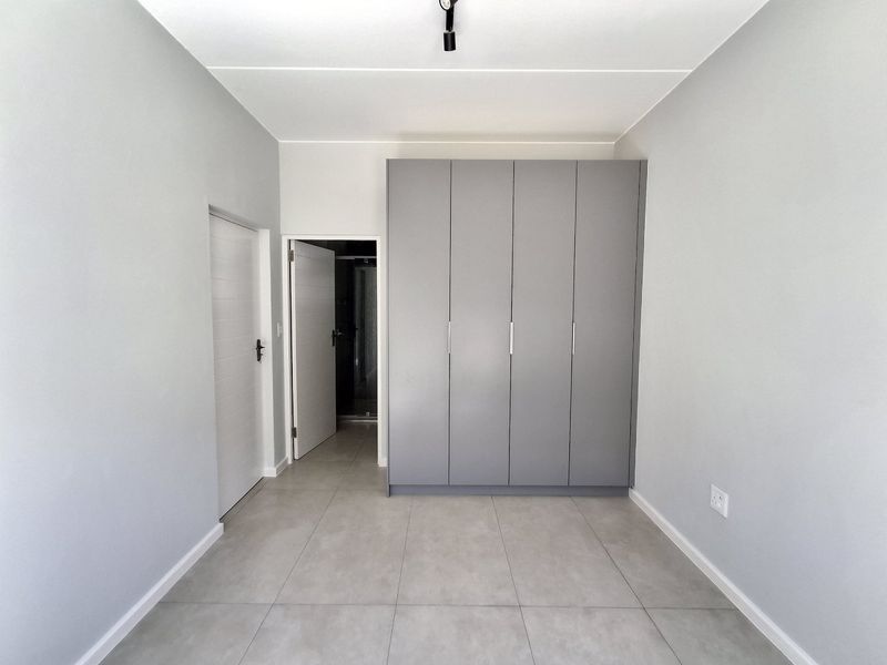 A clean, modern bedroom featuring neutral grey walls, large-format floor tiles, and ample built-in cupboard storage with a minimalist aesthetic.