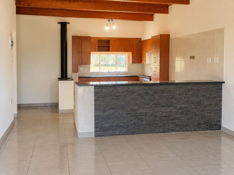 Spacious open-plan living and kitchen area featuring exposed timber ceiling beams, tiled flooring, a stone-clad breakfast bar, and a built-in fireplace.