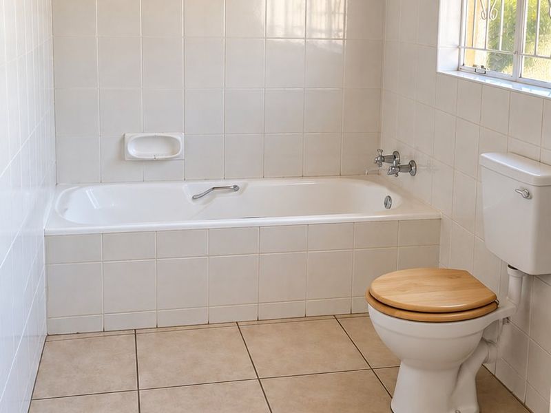 A functional bathroom featuring a built-in bathtub, ceramic tiling throughout, and a timber-paneled ceiling, offering a clean and neutral space.