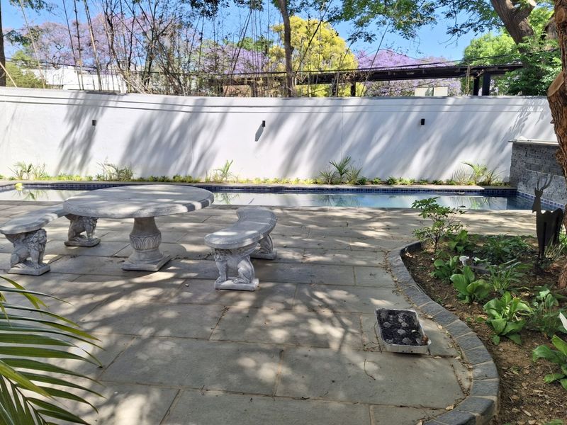A well-maintained outdoor pool area featuring stone paving, a swimming pool, and a perimeter boundary wall with established garden beds.