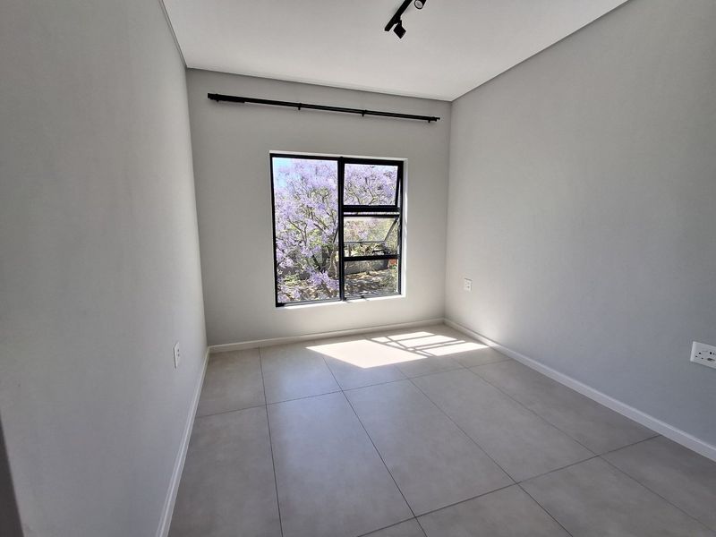 A bright, empty bedroom featuring modern tiled flooring, neutral walls, and a black aluminium window frame with a pleasant garden view.