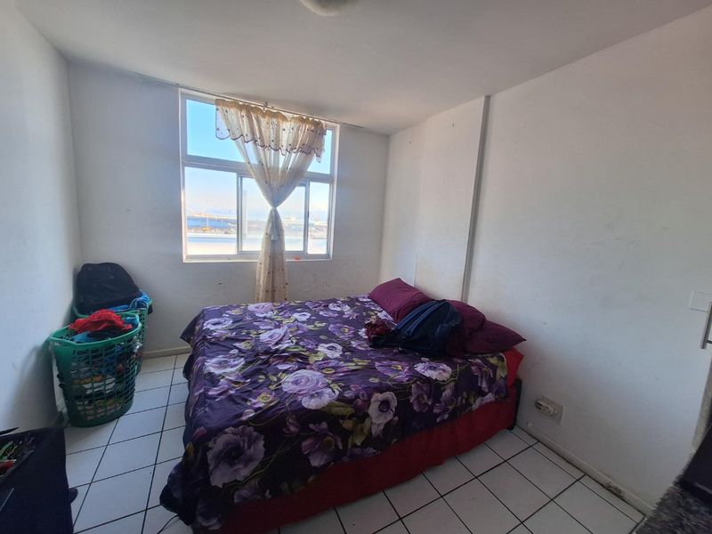 A bedroom featuring tiled flooring and a large window with natural light. The walls show signs of wear and require cosmetic attention.
