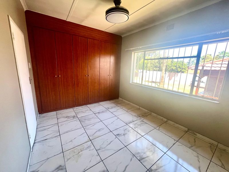 A bright, empty bedroom featuring built-in wooden cupboards, marble-look tiled flooring, and a window fitted with security bars.