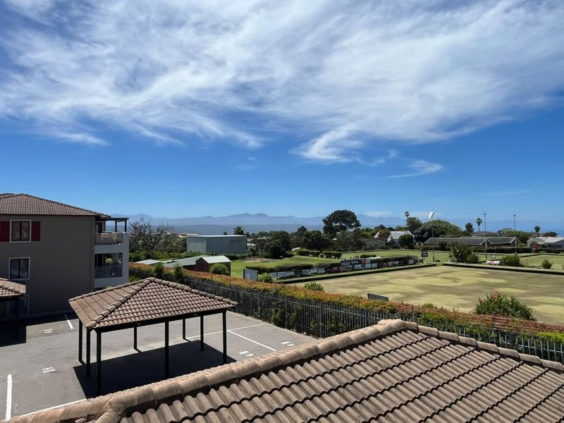 Elevated view overlooking a bowling green, surrounding residential area, and distant mountain range, featuring tiled roof structures and paved parking.