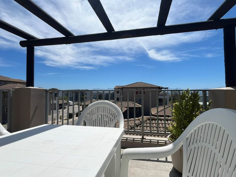 A sunny balcony featuring a pergola structure, metal balustrade, and tiled flooring with views over the surrounding residential complex.