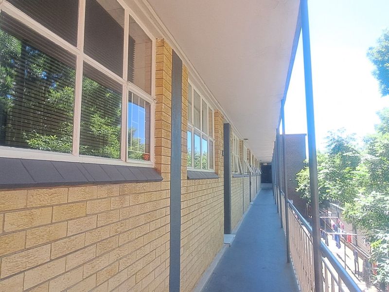 An exterior walkway or balcony featuring brick wall finishes, metal safety railings, and large multi-pane windows overlooking the surrounding trees.