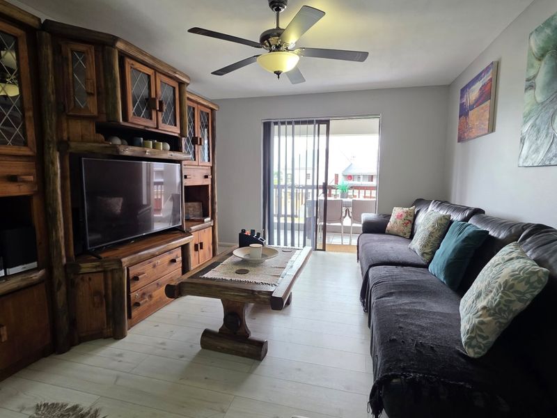 A comfortable living room featuring light-toned laminate flooring, a ceiling fan, and a sliding glass door providing access to an outdoor balcony area.