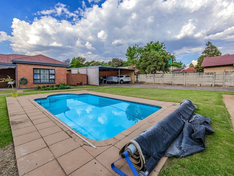 Spacious backyard featuring a swimming pool with paved surround, a covered carport, and a well-maintained lawn enclosed by a perimeter wall.