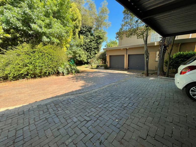 A paved driveway leading to a double garage with an attached carport, surrounded by established trees and garden greenery.