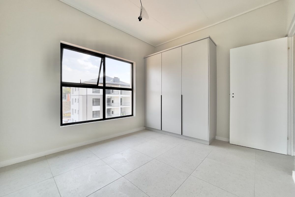 A bright, modern bedroom featuring neutral tiled flooring, a built-in wardrobe, and large aluminium-framed windows offering natural light.