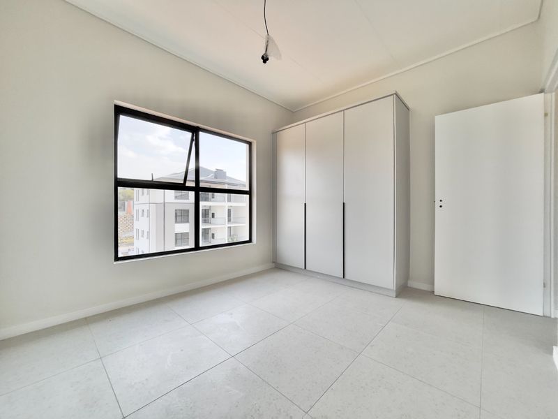 A bright, modern bedroom featuring neutral tiled flooring, a built-in wardrobe, and large aluminium-framed windows offering natural light.