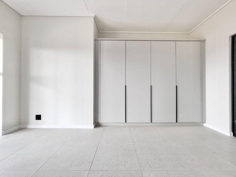A bright, modern bedroom featuring neutral tiled flooring and sleek, floor-to-ceiling built-in cupboards with minimalist handles.