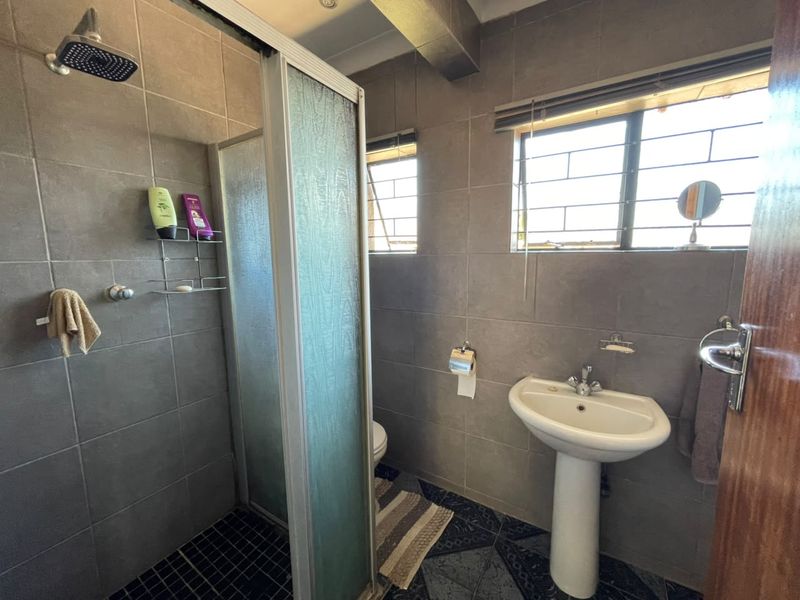 A functional bathroom featuring a shower enclosure, pedestal basin, and neutral wall tiling with natural light from two windows.
