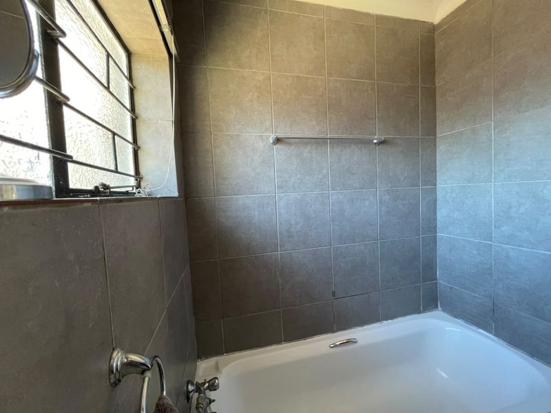 A functional bathroom featuring a built-in bathtub with grey wall tiling and a window with security bars.