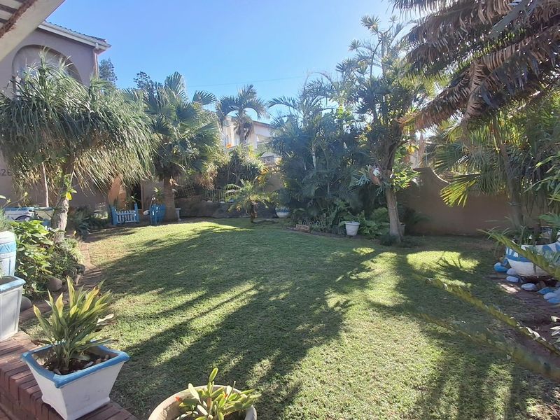 A lush, established garden featuring a well-maintained lawn, mature palm trees, and a brick-edged patio area.
