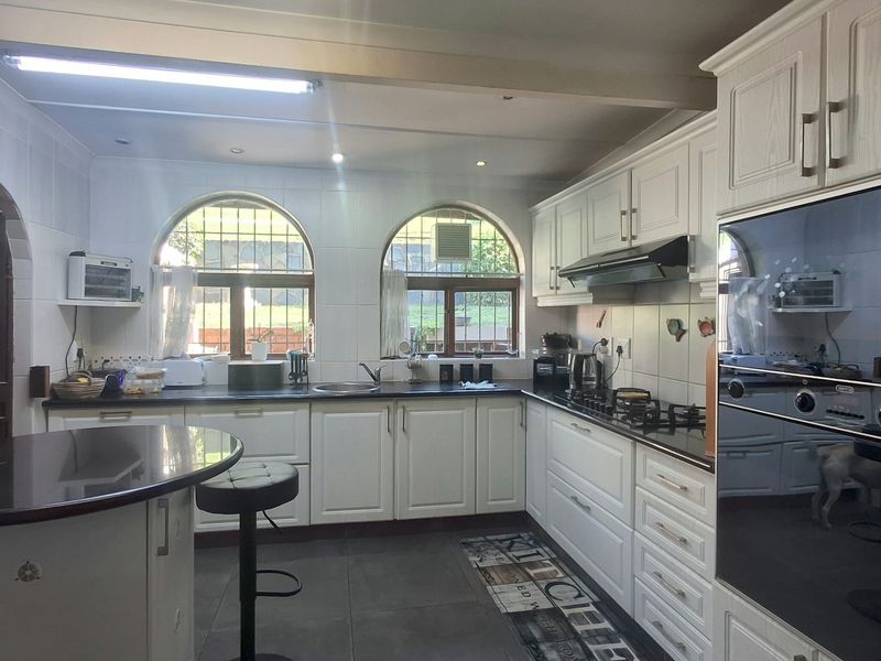 A well-lit kitchen featuring white cabinetry, dark stone countertops, a gas hob, built-in oven, and arched windows overlooking the garden.