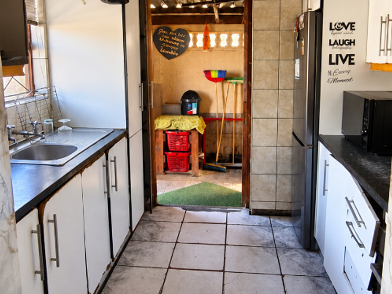 A galley-style kitchen featuring white cabinetry, tiled flooring, and a stainless steel sink, leading through to an outdoor utility area.
