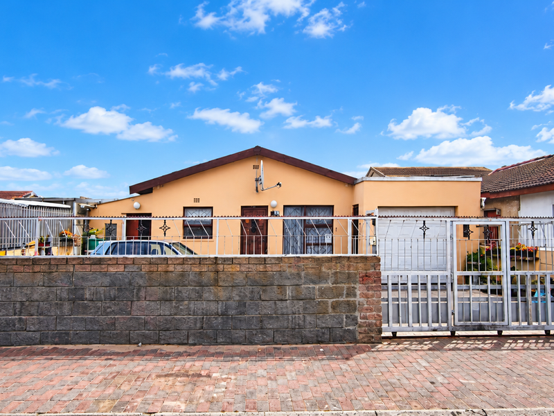 Front elevation of a single-story house featuring a brick boundary wall, metal security gate, paved driveway, and a tiled roof.