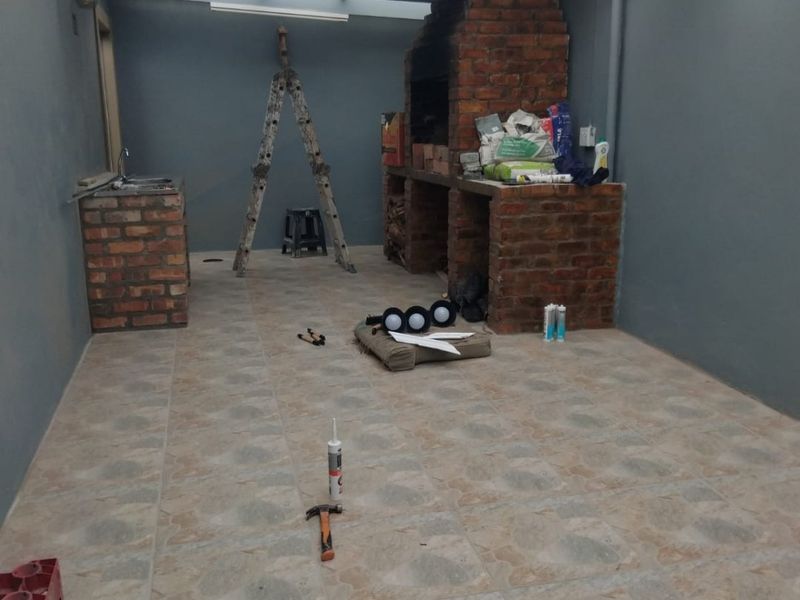 Covered outdoor patio area featuring a brick built-in braai, prep counter with sink, and tiled flooring, currently undergoing finishing works.