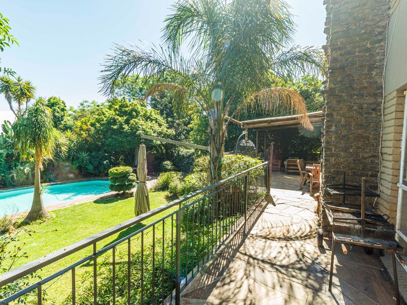 A covered stone-paved patio area overlooking a landscaped garden and swimming pool, featuring a brick exterior wall and metal safety railing.