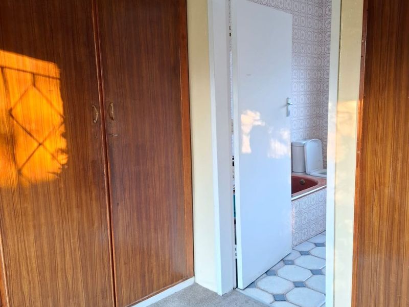 A hallway featuring built-in wooden cupboards and carpeted flooring, providing access to an adjacent bathroom with dated tiling.