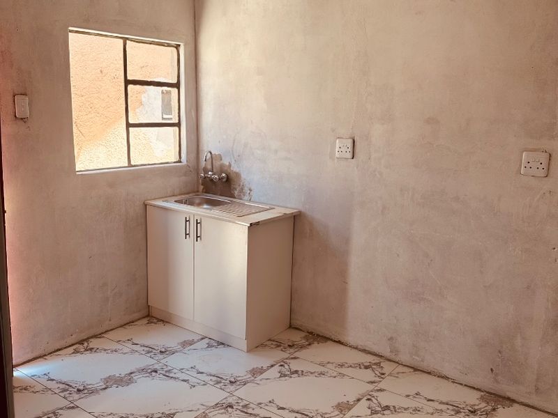 A compact, unfinished kitchen area featuring tiled flooring, a single basin sink with base cabinetry, and unpainted plaster walls.