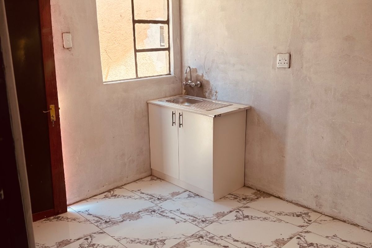 A compact, unfinished kitchen area featuring tiled flooring, a single basin sink with cabinet, and raw plaster walls awaiting final finishing.