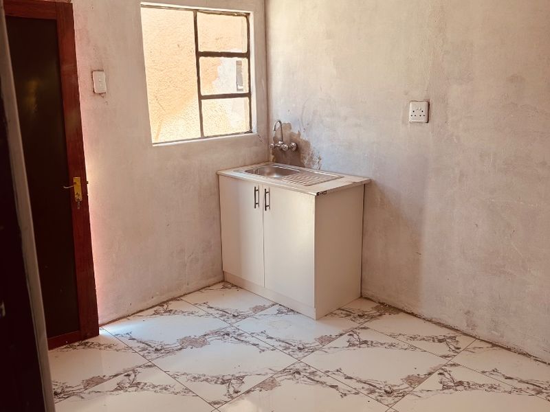A compact, unfinished kitchen area featuring tiled flooring, a single basin sink with cabinet, and raw plaster walls awaiting final finishing.