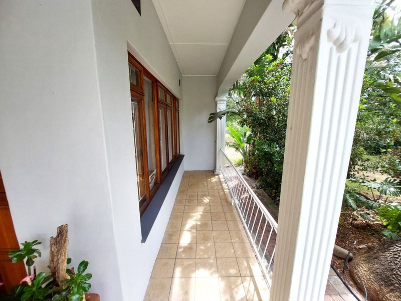 A covered patio area featuring tiled flooring, decorative columns, and a metal safety railing, overlooking the garden.