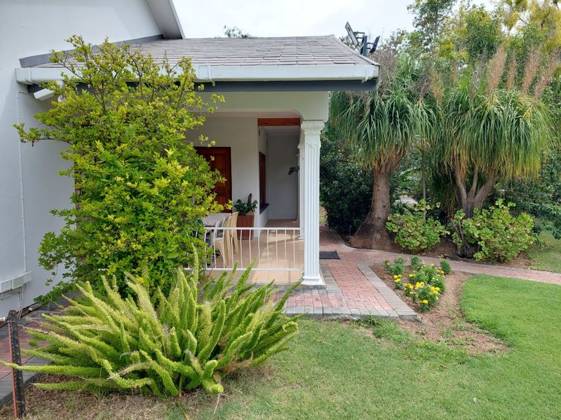 A covered patio area featuring tiled flooring, a decorative column, and a metal railing, overlooking a landscaped garden with a brick paved walkway.