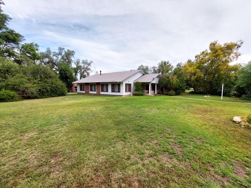 A spacious rear view of the house featuring a large, well-maintained lawn, mature garden surroundings, and a covered patio area.