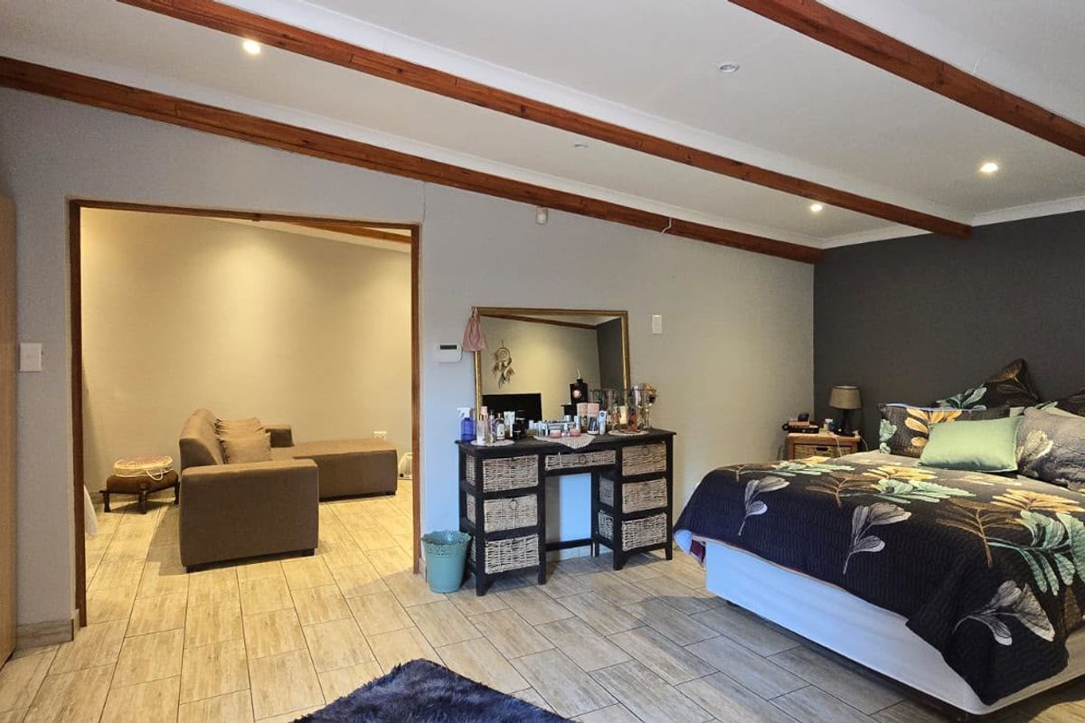 Spacious bedroom featuring exposed timber ceiling beams, tiled flooring, and a large built-in wardrobe, with an adjoining living area visible through the doorway.