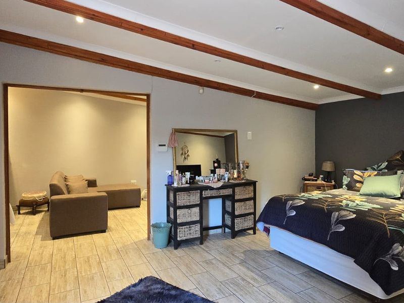 Spacious bedroom featuring exposed timber ceiling beams, tiled flooring, and a large built-in wardrobe, with an adjoining living area visible through the doorway.