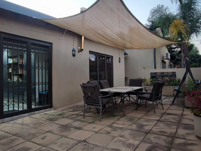 A paved outdoor patio area featuring a shade sail, sliding glass doors with security gates, and a brick boundary wall.
