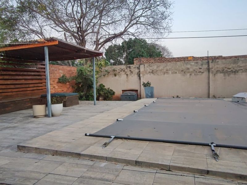 Outdoor pool area featuring a paved patio, timber pergola, built-in braai, and a covered swimming pool, enclosed by high brick boundary walls.