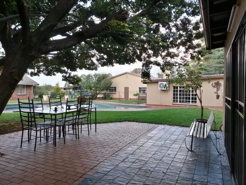 A spacious paved patio area overlooking a private swimming pool and well-maintained garden, featuring mature trees and secure perimeter fencing.