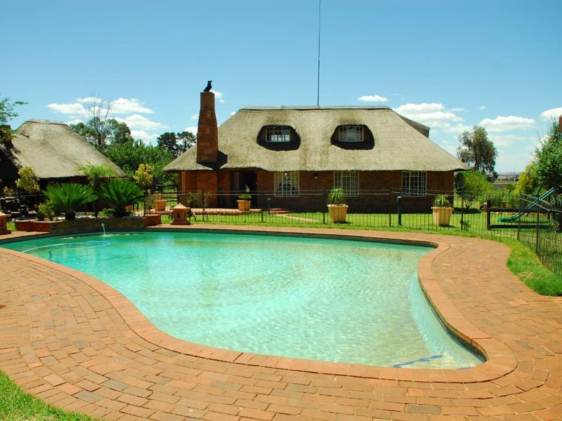 A well-maintained swimming pool area featuring brick paving, surrounded by a landscaped garden and a traditional thatch-roofed farmhouse in the background.