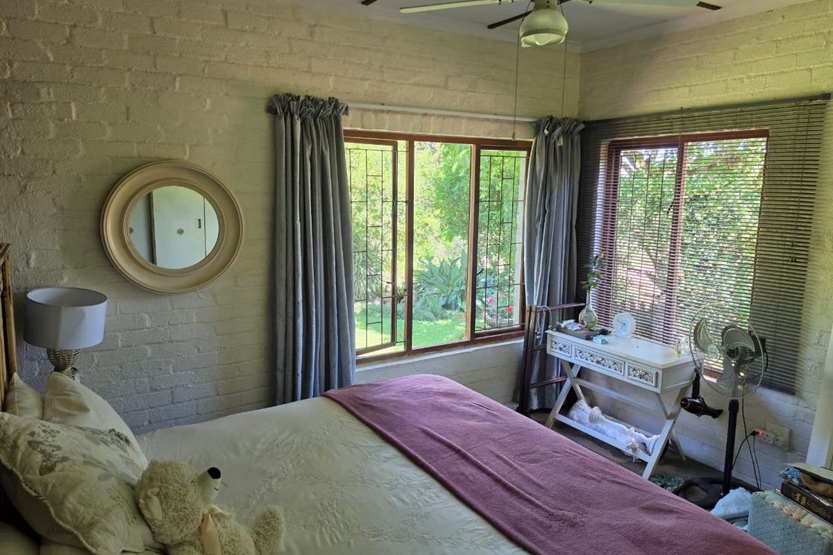 A cozy bedroom featuring exposed brick walls, a ceiling fan, and large timber-framed windows with security bars overlooking the garden.