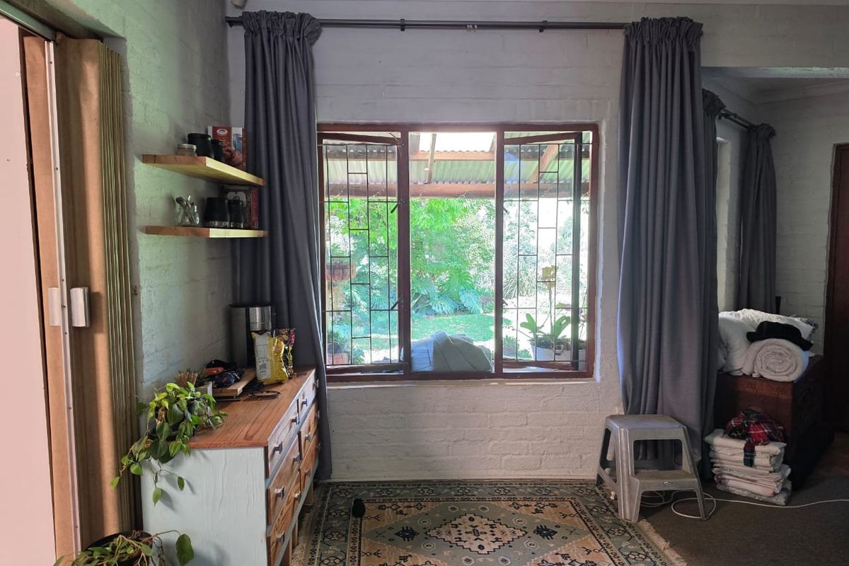 A living area featuring exposed brick walls, timber-framed windows with security bars, and floating timber shelving, offering a view into the garden.
