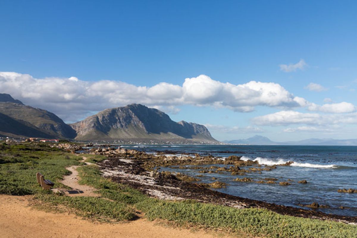 A scenic coastal view featuring a rugged shoreline, natural vegetation, and distant mountain ranges under a clear blue sky.