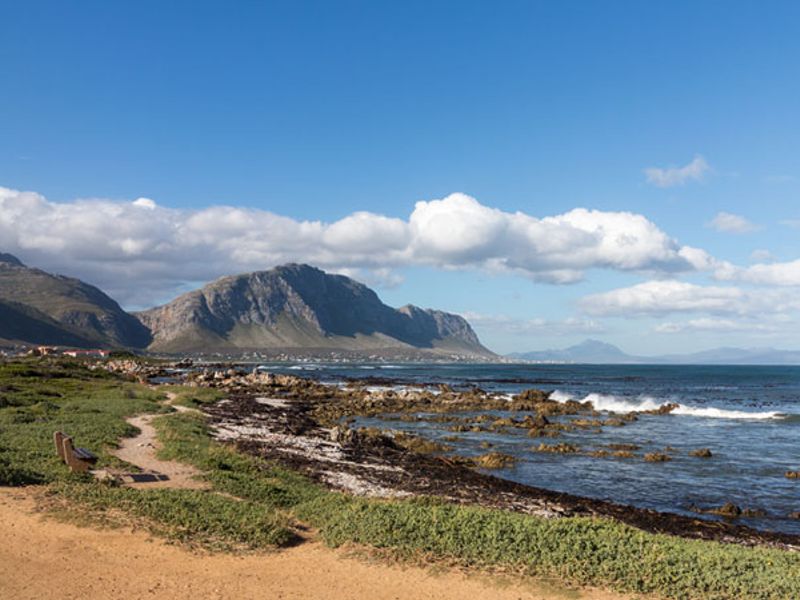 A scenic coastal view featuring a rugged shoreline, natural vegetation, and distant mountain ranges under a clear blue sky.