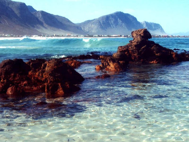 A scenic coastal view featuring rocky outcrops, clear shallow water, and distant mountain ranges.
