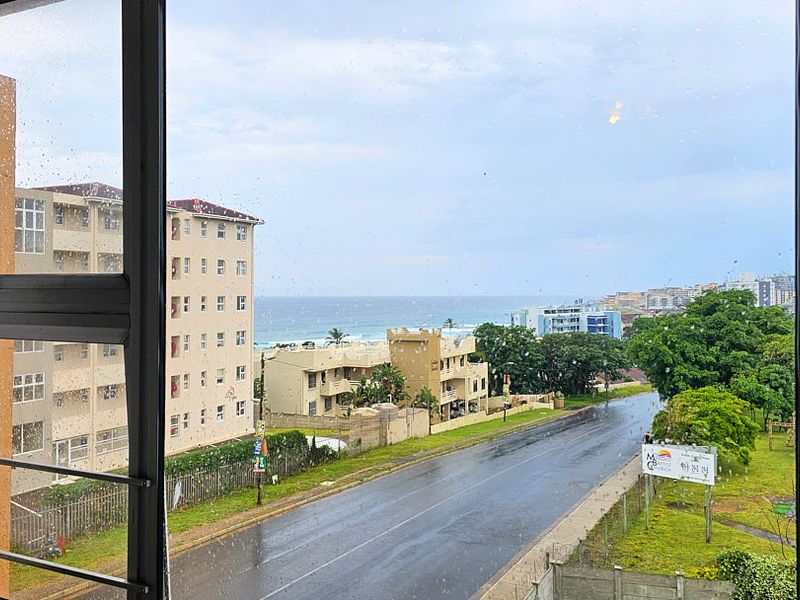 A view from an apartment window overlooking a street, surrounding residential buildings, and the ocean in the distance.